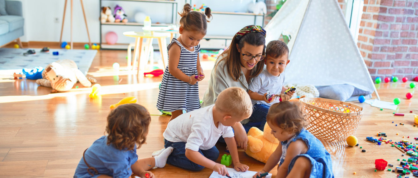 Kindergartenpädagogin sitzt mit Papier und Bleistift auf dem Boden und spielt im Kindergarten mit den Kindern mit Spielzeug.