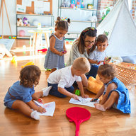 Kindergartenpädagogin sitzt mit Papier und Bleistift auf dem Boden und spielt im Kindergarten mit den Kindern mit Spielzeug.