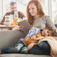 Kind kuschelt sich an lesende Mutter, Vater frühstückt entspannt