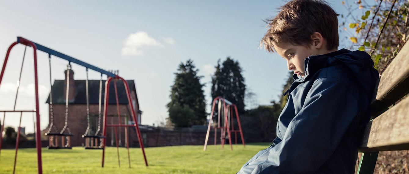 Kleiner Bub sitzt alleine auf Bank am Spielplatz