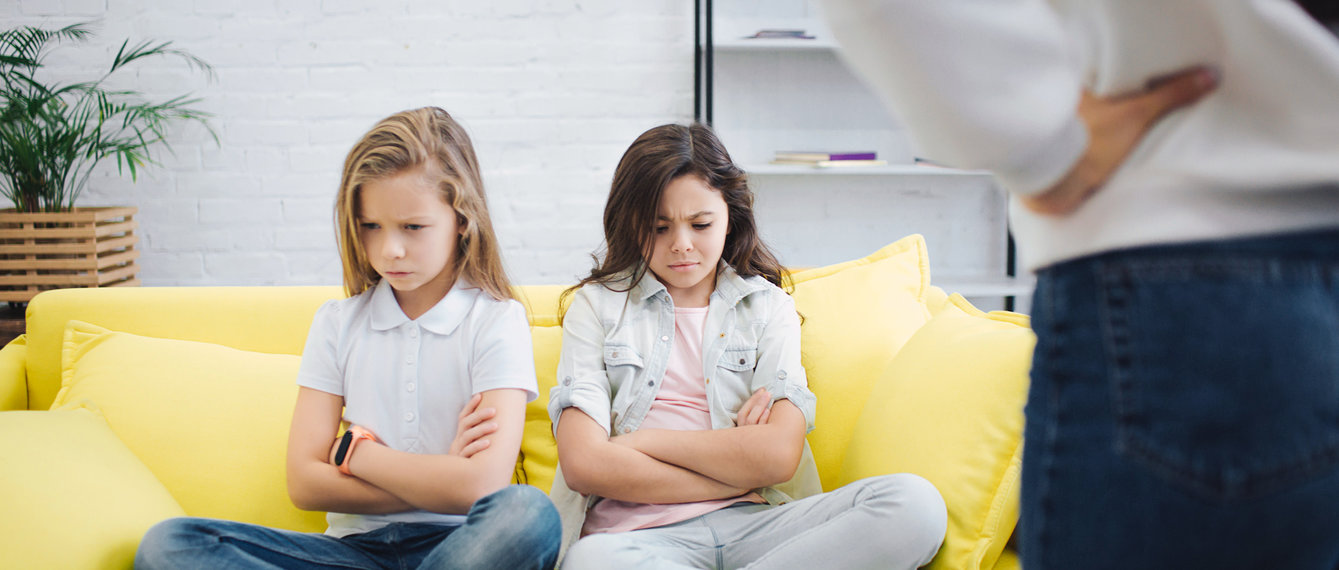 Traurige und verärgerte Schulkinder sitzen auf Sofa im Zimmer und schauen verärgert auf den Boden. Halten sie Hände und Beine gekreuzt. Junge Frau steht vor ihnen. Sie halten die Hände auf den Hüften.