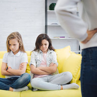 Traurige und verärgerte Schulkinder sitzen auf Sofa im Zimmer und schauen verärgert auf den Boden. Halten sie Hände und Beine gekreuzt. Junge Frau steht vor ihnen. Sie halten die Hände auf den Hüften.