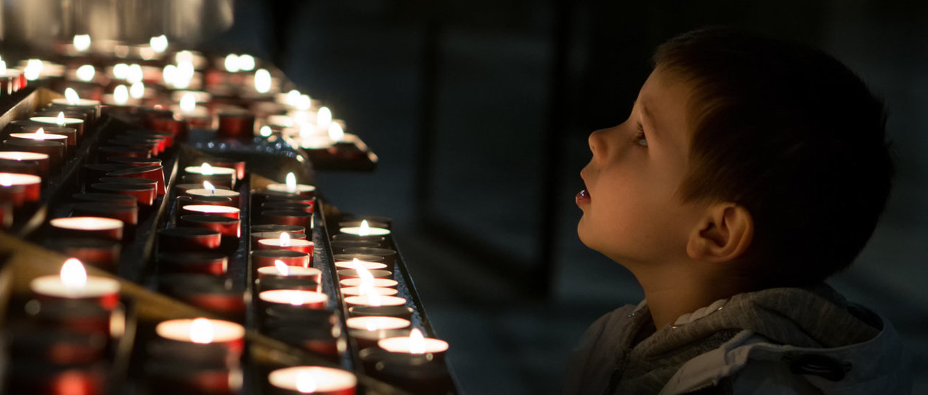 Kleiner Bub in dunkler Kirche vor Teelichtern, die angezündet sind, von Seite zu sehen