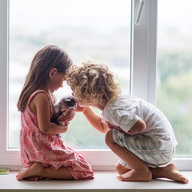 zwei Kinder sitzen vorm Fenster und kuscheln mit einer Katze
