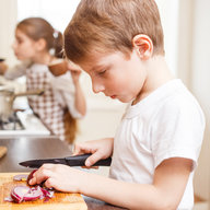 Junge schneidet Zwiebel und Gemüse, im Hintergrund ein Mädchen, beide in der Küche