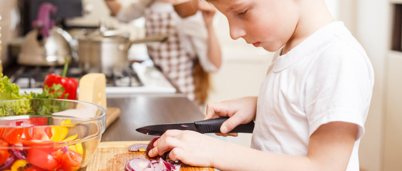 Junge schneidet Zwiebel und Gemüse, im Hintergrund ein Mädchen, beide in der Küche